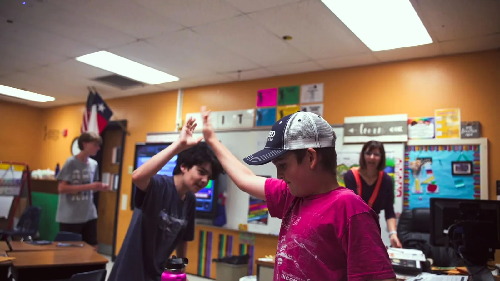 Two boys high-five in a sunny classroom while their teacher works in the background